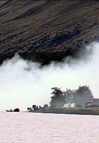 A boat rests near the shore of a lake backed by rolling clouds and a towering hill.